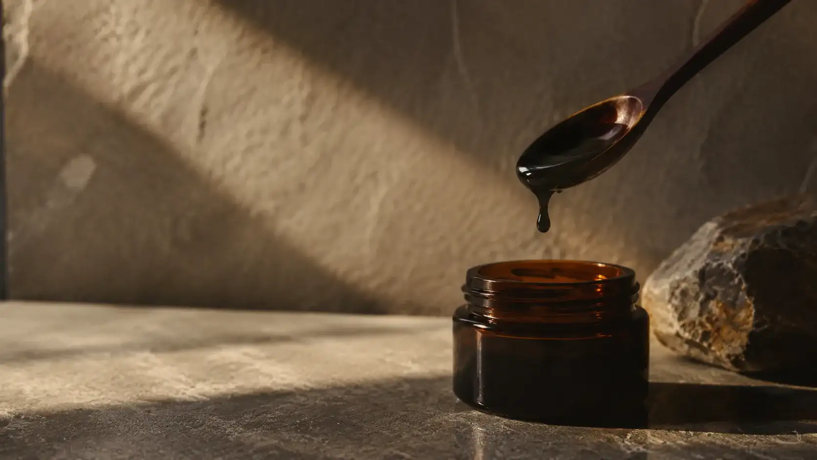 A single dark resin drop suspended from a spoon above a glass jar in long low morning light.