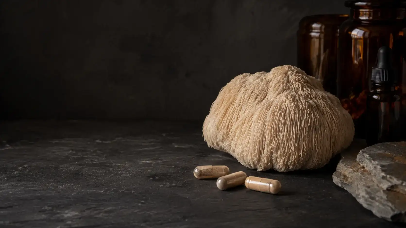 A whole dried lion's mane fruiting body, pale and shaggy, lying on dark slate beside three unbranded capsules in cold north light.