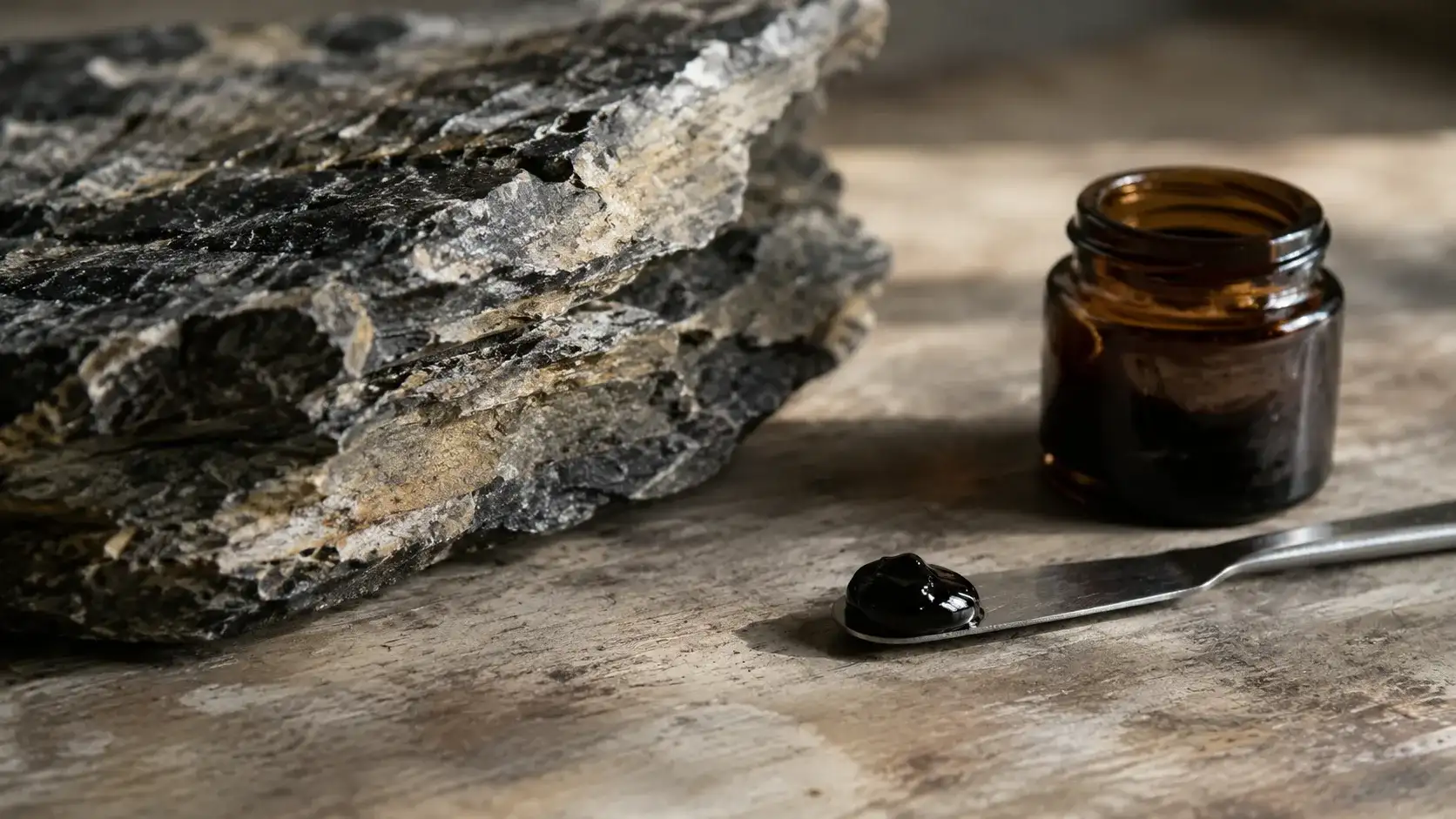 A bead of near-black shilajit resin on a small steel spatula beside layered mineral rock under raked daylight.