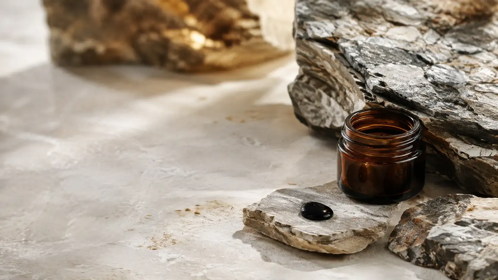 A single bead of near-black shilajit resin resting on pale mineral stone beside a fissured slab of rock in cold daylight.