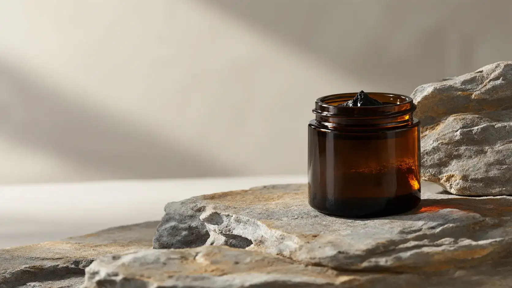 Daylight passing through an unbranded amber glass jar of dark shilajit resin standing on pale layered mineral rock.