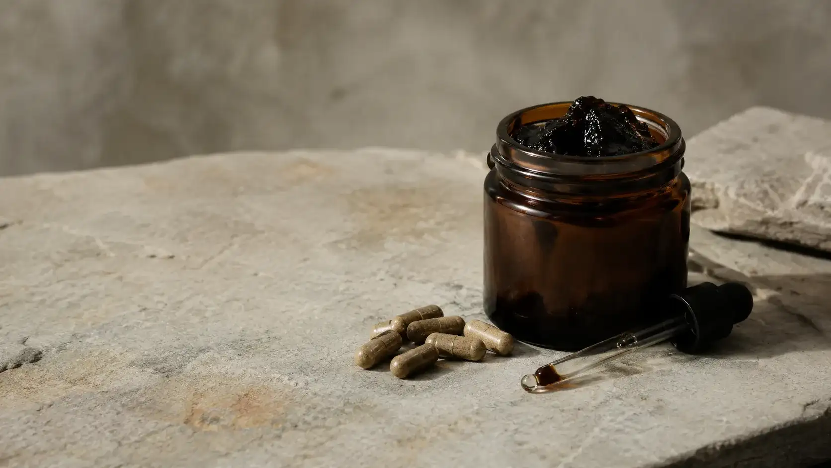 An unbranded amber jar of dark resin, a small pile of plain capsules and a glass dropper arranged on pale stone in even daylight.