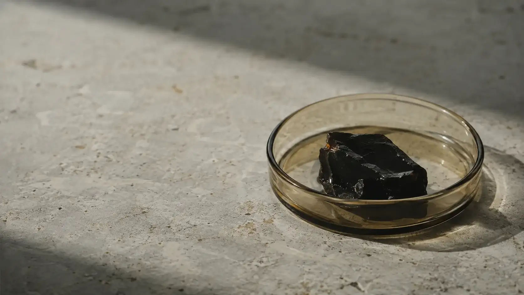 A single dark resin sample in a plain glass dish on a pale stone surface under flat north light.