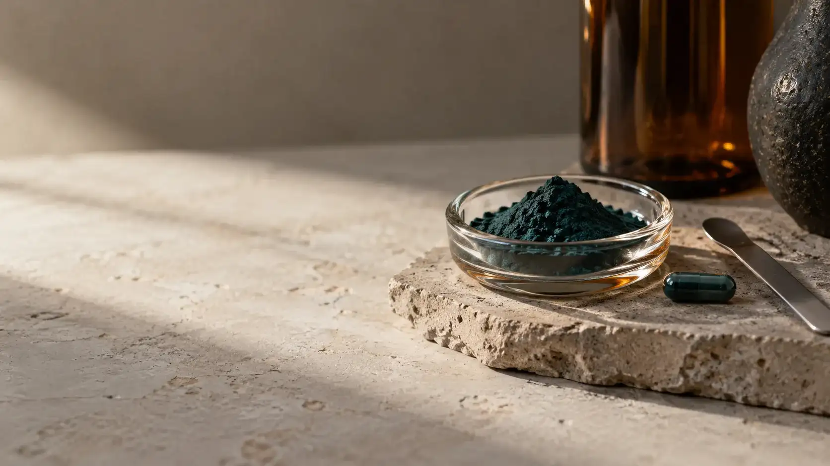 A small glass dish of deep blue-green spirulina powder on pale stone beside a steel spatula and one unbranded capsule in north light.